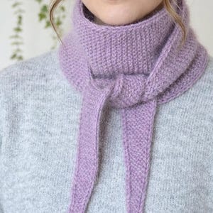 May include: A soft, lavender-colored knitted scarf with a textured, ribbed pattern. The scarf is wrapped around the neck and tied in a simple knot at the front, with two long, tapered ends hanging down over a light gray, textured knit sweater.