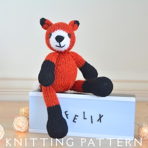 May include: A knitted fox toy in orange, black, and white, with the name "FELIX" on a light box. The fox has a round body, black paws, and a white face. The text "KNITTING PATTERN" is at the bottom.