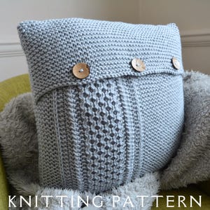 以下が含まれることがあります： ケーブル編みのパターンと3つの木製ボタンが付いたグレーのニット枕。枕はふわふわの薄い茶色の毛布の上に置かれています。