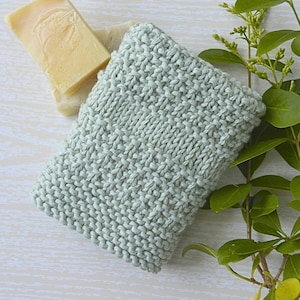 May include: A rectangular, light sage green knitted washcloth with a textured stitch pattern, positioned next to two stacked rectangular bars of pale yellow soap and a green leafy branch with small buds on a light, wood-grain surface.