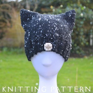 May include: A dark gray knit hat with cat ears, speckled with white flecks, sits on a mannequin head. The hat has a small white tag with the text "THE WOOLLY DESIGN CO." The words "KNITTING PATTERN" are at the bottom.