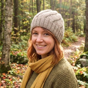 Pode incluir: Uma mulher usando um gorro de malha bege, um cachecol amarelo mostarda e um suéter verde. O gorro tem um padrão texturizado. O fundo é uma floresta com árvores e folhas caídas.