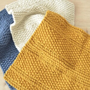 May include: Three square knitted cloths in solid colours, featuring a textured garter stitch pattern. The set includes one mustard yellow cloth, one cream white cloth, and one slate blue cloth, all arranged in a slightly overlapping layered stack.