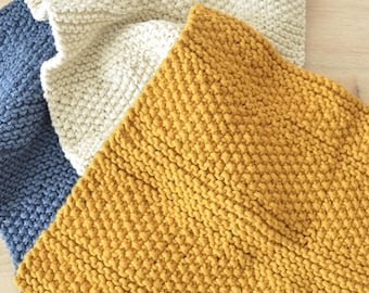 May include: Three square knitted cloths in solid colors, featuring a textured garter stitch pattern. The set includes one mustard yellow cloth, one cream white cloth, and one slate blue cloth, all arranged in a slightly overlapping layered stack.