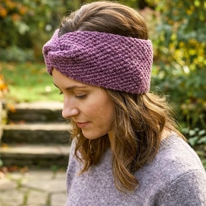 Puede incluir: Una diadema morada tejida con un diseño de nudo retorcido. La diadema está hecha de un hilo grueso y texturizado y se usa en la cabeza. La persona de la imagen lleva un suéter gris. El fondo está borroso con tonos verdes y marrones.