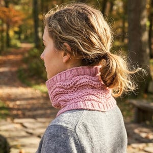 May include: A pink knitted cowl neck warmer. The cowl is a soft pink color and has a textured, ribbed design. The person is wearing a gray sweater. The background is a blurred view of a forest with fall foliage.