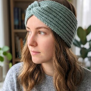 May include: A knitted headband in a soft, muted green color. The headband has a twisted knot detail at the top and a ribbed texture. The accessory is made of a soft, possibly wool, material.
