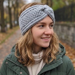 May include: A gray, hand-knitted headband with a twisted knot design. The headband is made of a textured yarn and is worn across the forehead. This winter accessory is perfect for keeping warm during cold weather.