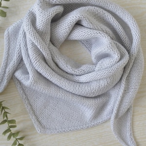 May include: A light gray triangular scarf featuring a consistent garter stitch knit pattern. The scarf is folded into a soft, draped shape with pointed ends, showcasing the uniform texture of the light gray yarn throughout the entire accessory.