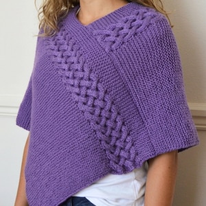May include: A knitted purple poncho with a V-neck design. The poncho features a cable knit pattern along the shoulder and front. The short-sleeved garment is made of a soft, textured yarn.