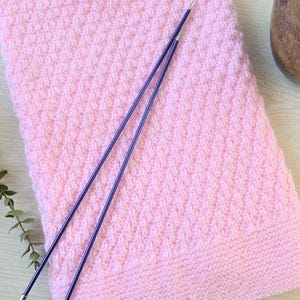 May include: A soft pink knitted blanket with a textured diamond pattern. Two long, thin, purple knitting needles rest on the blanket. The blanket is folded and appears to be made of a soft yarn. The needles are made of metal.