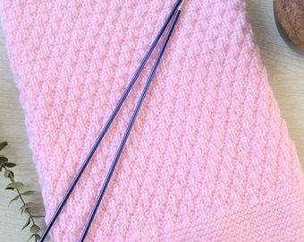 Könnte beinhalten: Eine hellrosa gestrickte Decke mit einem strukturierten Rautenmuster. Zwei lange, dünne, violette Stricknadeln liegen auf der Decke. Die Decke ist gefaltet und scheint aus weichem Garn zu bestehen. Die Nadeln sind aus Metall.