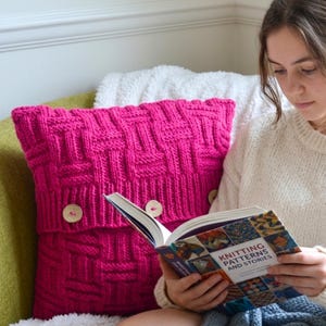 Könnte beinhalten: Ein leuchtend pinkfarbenes Strickkissen mit einem strukturierten Muster und Holzknöpfen. Das Kissen liegt auf einem grünen Stuhl mit einer weißen, flauschigen Decke. Ein Buch mit dem Titel "Knitting Patterns and Stories" wird gelesen.