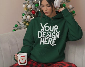 May include: A dark green hoodie with the text "YOUR DESIGN HERE" in white. The person is wearing red and black plaid pajama pants and holding a mug with a Santa Claus design. A Christmas tree is in the background.