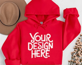 Könnte beinhalten: Ein leuchtend roter Kapuzenpullover mit Kapuze und Vordertasche. Der Kapuzenpullover zeigt den Text "YOUR DESIGN HERE" in einer weißen, abgenutzten Schriftart. Ein beiger Hut ist im Hintergrund zu sehen.
