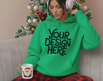 May include: A person wearing a green hoodie with the text "YOUR DESIGN HERE" in black. The person is wearing white headphones and holding a mug with a Santa Claus design. The person is wearing red and black plaid pajama pants. A Christmas tree is in the background.