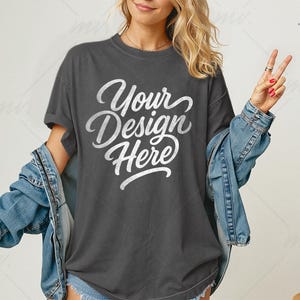 May include: Dark gray t-shirt with the text "Your Design Here" in a white script font. A light blue denim jacket is draped over the shoulders. The shirt is a relaxed fit, and the model is wearing a ring.