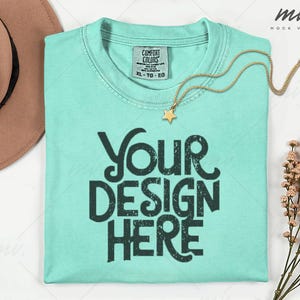 May include: A light turquoise t-shirt with the text "YOUR DESIGN HERE" in dark green. A gold necklace with a star pendant rests on the shirt. A brown hat is visible in the upper left corner.