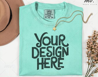 May include: A light turquoise t-shirt with the text "YOUR DESIGN HERE" in dark green. A gold necklace with a star pendant rests on the shirt. A brown hat is visible in the upper left corner.