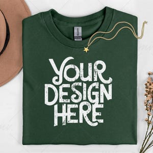 May include: A dark green t-shirt with the text "YOUR DESIGN HERE" in distressed white lettering. A gold necklace with a star pendant is draped across the shirt. A brown hat and dried flowers are in the background.