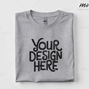 May include: A folded light gray t-shirt with the text "YOUR DESIGN HERE" in a distressed black font. The shirt has a crew neck and a tag is visible on the collar.