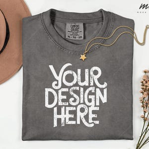 May include: A gray t-shirt with the text "YOUR DESIGN HERE" in white. A gold necklace with a star pendant rests on the shirt. A brown hat is visible in the upper left corner. The shirt has a Comfort Colors tag.