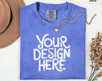 May include: A periwinkle blue t-shirt with the text "YOUR DESIGN HERE" in white. A gold necklace with a star pendant rests on the shirt. A brown hat is in the upper left corner. The shirt has a Comfort Colors tag.