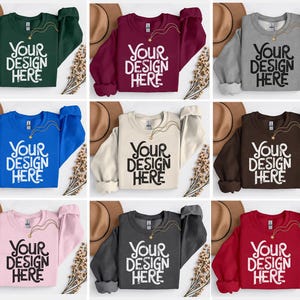 Puede incluir: Una colección de sudaderas de cuello redondo en varios colores: verde, burdeos, gris, azul, beige, marrón, rosa, carbón y rojo. Cada sudadera presenta el texto "YOUR DESIGN HERE" en una fuente audaz y estilizada.