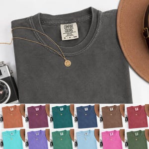 May include: A charcoal gray crew neck t-shirt with a Comfort Colors label, featuring a gold-toned chain necklace with a small smiley face pendant. The shirt is displayed alongside a brown hat and a vintage-style camera, with various color options.