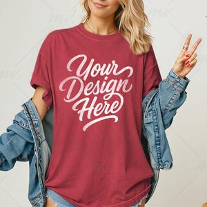 May include: A maroon t-shirt with the text "Your Design Here" in a white script font. The model is wearing a denim jacket over the shirt. The background is a neutral color with a decorative plant in a woven basket.