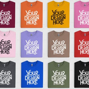 Puede incluir: Una colección de camisetas plegadas en varios colores, incluyendo burdeos, naranja, amarillo, rosa, rosa claro, morado, marrón, rojo, gris oscuro, azul, verde oliva y negro. Cada camiseta muestra el texto "YOUR DESIGN HERE" en blanco.