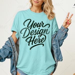 May include: A light blue t-shirt with the text "Your Design Here" in a black script font. The person is wearing a denim jacket over the shirt. The image is taken in a studio setting with a neutral background.
