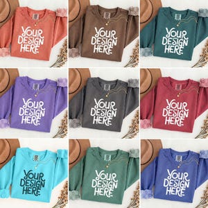 May include: A collection of nine crew neck sweatshirts in various colors, including rust, brown, teal, purple, gray, burgundy, turquoise, green, and blue. Each sweatshirt features the text "YOUR DESIGN HERE" in white block letters.