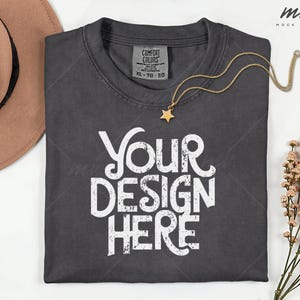May include: A dark gray t-shirt with the text "YOUR DESIGN HERE" in white, a gold star pendant necklace, and a Comfort Colors tag. The shirt is folded neatly on a white surface.