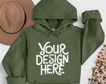 May include: An olive green hoodie with the text "YOUR DESIGN HERE" in white. The hoodie has a drawstring hood, a front pocket, and rolled-up sleeves. A brown hat is visible in the background.