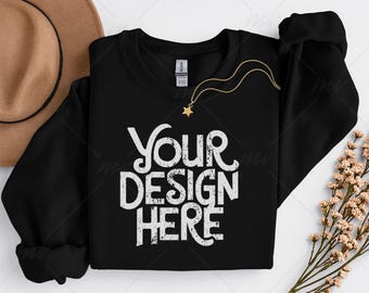 May include: A black crewneck sweatshirt with the text "YOUR DESIGN HERE" in white, a gold necklace with a star pendant, and a brown hat. The sweatshirt is folded, with a sprig of flowers on the side.