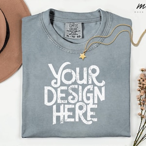 May include: A light blue t-shirt with the text "YOUR DESIGN HERE" in white. A gold necklace with a star pendant rests on the shirt. A brown hat and sprigs of dried flowers are also in the image.