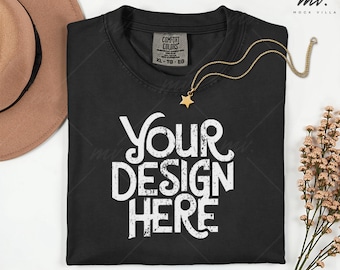 May include: Black t-shirt with the text "Your Design Here" in white, featuring a gold star pendant necklace. The shirt has a Comfort Colors tag and is laid flat on a white surface. A tan hat and flowers are in the background.