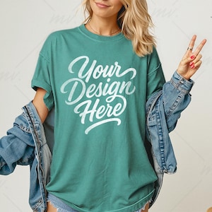 Op de afbeelding: Tealkleurig T-shirt met de tekst "Your Design Here" in witte letters. Een spijkerjasje is over de schouders gedrapeerd. De persoon draagt een ring en maakt het vredesteken.