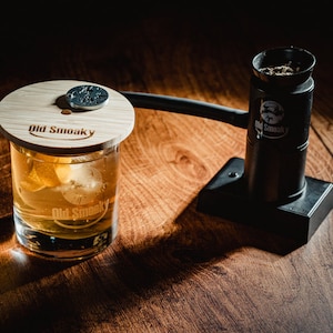 May include: A glass of whisky with ice, topped with a wooden lid featuring the text "Old Smoky" and a metal coin. A black metal device with the text "Old Smoky" is also visible.