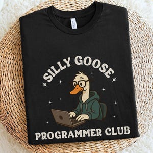 May include: Black t-shirt with the words "SILLY GOOSE PROGRAMMER CLUB" in white. The graphic features a cartoon goose wearing glasses and a hoodie, working on a laptop. The shirt is displayed on a woven surface.