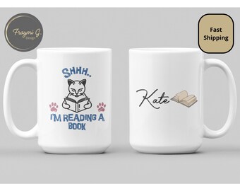 May include: White ceramic mug with a black and white illustration of a cat reading a book. The text "Shhh... I'm reading a book" is printed in blue on the mug. The other mug is white with the name "Kate" written in black cursive and a brown open book illustration.