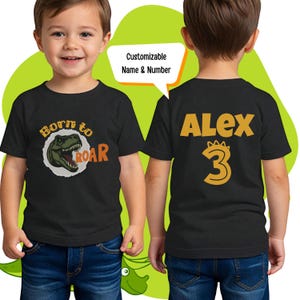 Peut inclure: T-shirt noir avec un motif de dinosaure et le texte "Born to ROAR". Le dos du t-shirt porte un nom et un numéro, "Alex 3", en orange.