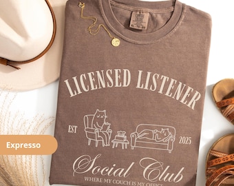 May include: Espresso-colored t-shirt with the text "LICENSED LISTENER Social Club" and a graphic of cats on a couch. The shirt is styled with a gold necklace, a beige hat, and brown sandals.