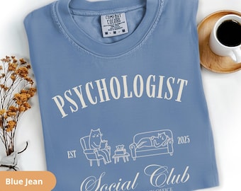 May include: Blue jean coloured t-shirt with the text "PSYCHOLOGIST Social Club" and a graphic of a cat and a person on a couch. The shirt also has the text "WHERE MY COUCH IS MY OFFICE" and "EST 2025".