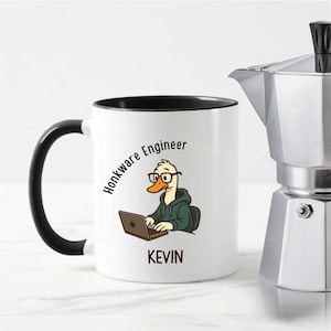 May include: White ceramic mug with a black handle and interior. The mug features a cartoon duck wearing glasses and a hoodie, working on a laptop. Text on the mug reads "Honkware Engineer" and "KEVIN". A silver coffee maker is in the background.