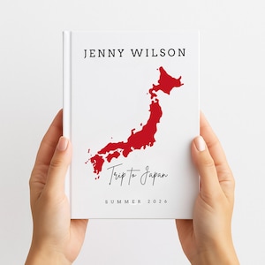 May include: A white journal with the text "JENNY WILSON" at the top and a red map of Japan. Below the map, the words "Trip to Japan" are written in cursive, with "SUMMER 2026" at the bottom. The journal is held by two hands.