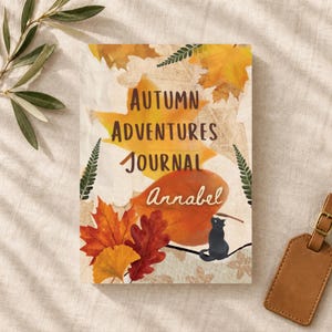 Peut inclure: Un journal rectangulaire avec un fond crème orné de feuilles d'automne orange, jaunes et rouges, de fougères vertes et d'un petit chat noir. Le texte indique Autumn Adventures Journal Annabel en écriture brune.