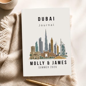 Könnte beinhalten: Ein weißes Notizbuch mit dem Text "DUBAI Journal" oben. Darunter befindet sich eine farbenfrohe Illustration der Skyline von Dubai. Das Notizbuch enthält auch die Namen "MOLLY & JAMES" und das Datum "SOMMER 2026".
