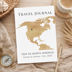 May include: A white travel journal with the text "TRAVEL JOURNAL" at the top and a gold-colored map of North America. The journal also includes the text "TRIP TO NORTH AMERICA Emma & James • Est. 2023". A compass, pen, and coffee cup are also visible.